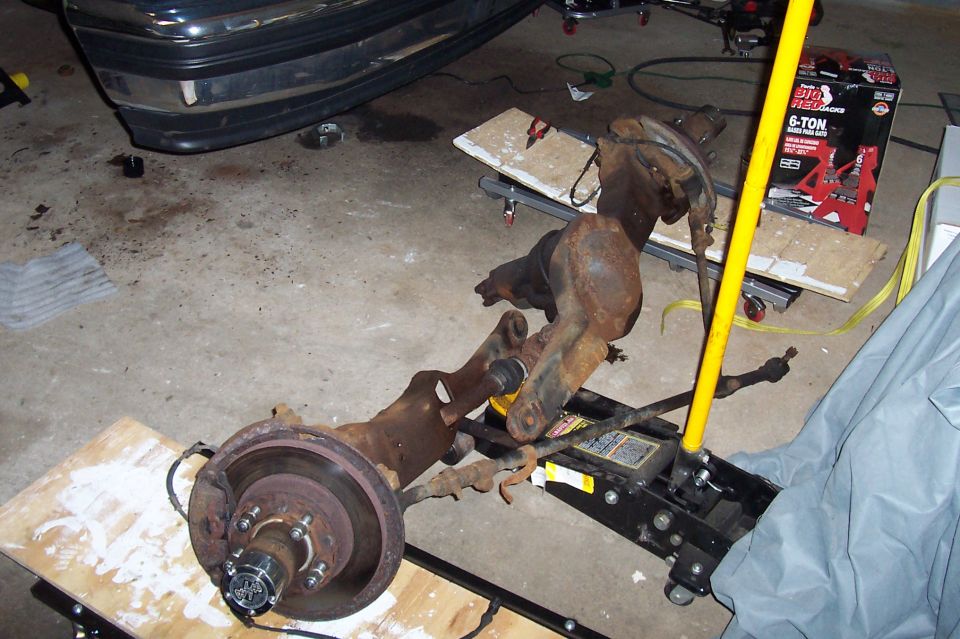 Rusty removed front axle with disc brake and locking hub on plywood and a floor jack in a garage