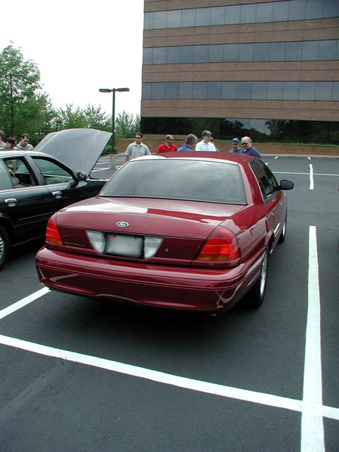 2002 Ford Crown Victoria - A group of people gather around vehicles in a parking lot, with a modern office building in the background.