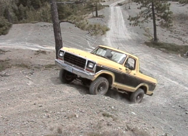 Yellow and black Ford Bronco climbing a dirt hill, front-left wheel elevated