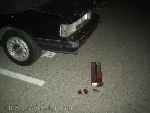 1985 Ford Mustang - A black car's rear end is damaged, with detached taillight components scattered on the asphalt.