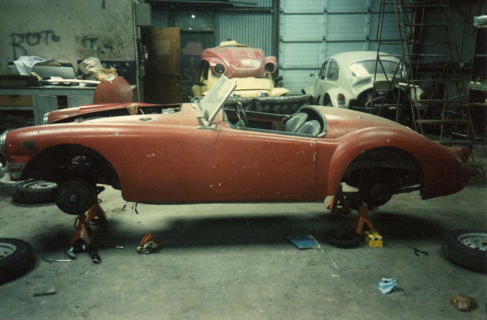 Red MGA roadster stripped and on jack stands in a restoration shop with VW Beetles behind