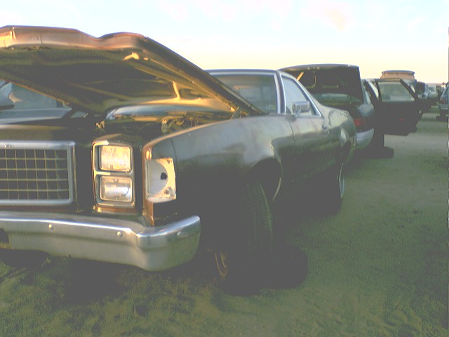Dark coupe with hood up, parked in a lot with other cars. Missing headlight