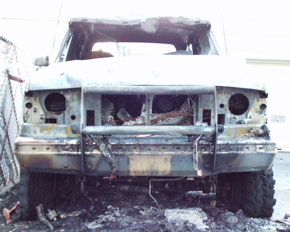Front view of a heavily damaged, burnt SUV. The grille, headlights, and much of the front end are destroyed, revealing internal components. The windshield and roof are also severely burned
