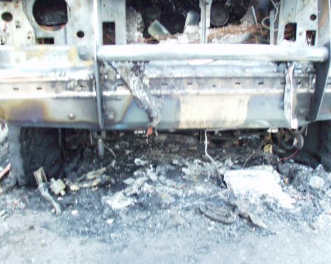1989 Ford Bronco - The front end of a 1989 Ford Bronco is heavily damaged by fire, with the grille and bumper melted and debris scattered on the ground.
