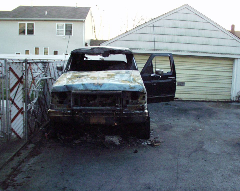 Burnt-out truck with open driver's door parked in front of a garage