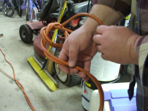1965 Ford Mustang - A person is holding an orange extension cord that is coiled up. In the background, there is a red riding mower and a yellow broom.