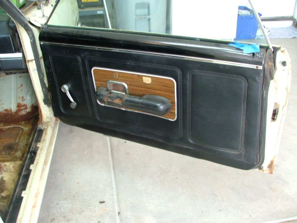 Black car door interior panel with chrome crank and wood-grain armrest, open in a garage