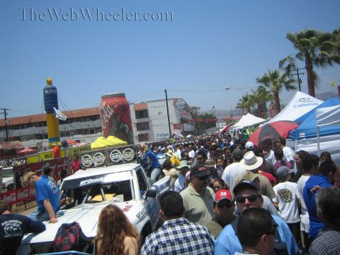 1990 Ford Bronco - large crowd fills a street between vendor booths and banners at a sunny outdoor festival, with an off-road race truck at left.