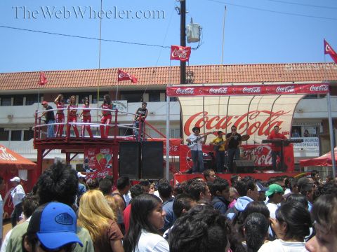1990 Ford Bronco - crowd watches performers on a red Coca-Cola sponsored stage at a sunny outdoor event near a tile-roofed building.
