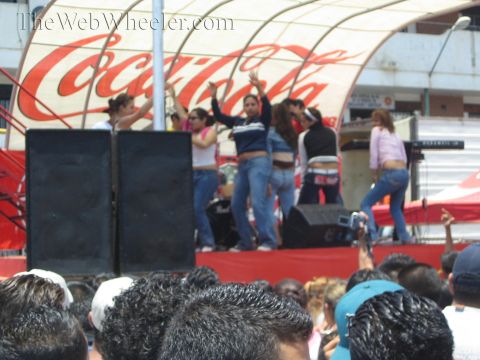 1990 Ford Bronco - People dance with raised arms on a stage in front of a red Coca-Cola backdrop while a crowd watches below.