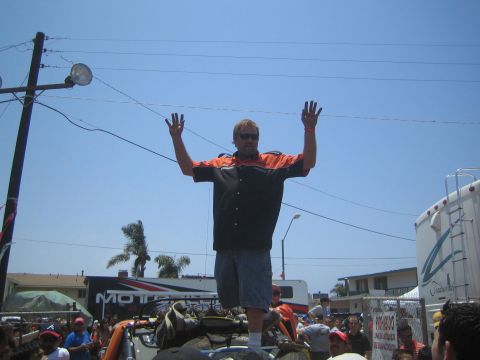 1990 Ford Bronco - man in an orange and black shirt and sunglasses stands with arms raised on top of a vehicle above an outdoor crowd.