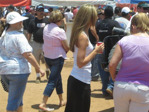 1990 Ford Bronco - crowd of people stands and mingles on dusty ground at a sunny outdoor event, one woman holding a red drink can.