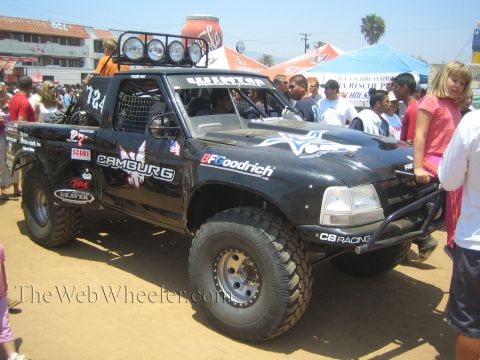 1990 Ford Bronco - black off-road prerunner race truck numbered 784, covered in sponsor decals with a roof-mounted light bar, sits amid a street crowd.