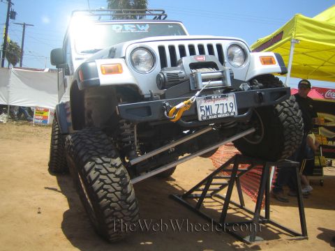 1990 Ford Bronco - white lifted Jeep Wrangler with a front winch and oversized mud tires is parked atop metal display ramps under a sunny sky.