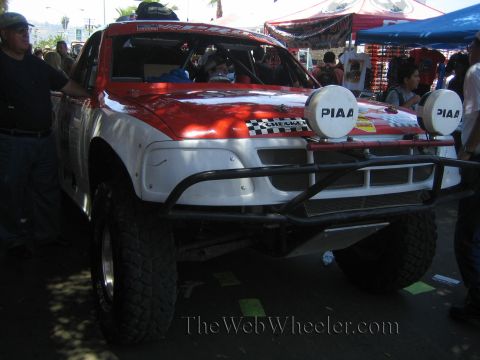 1990 Ford Bronco - red-and-white off-road race truck fitted with twin PIAA driving lights sits among vendors and spectators on a busy street.
