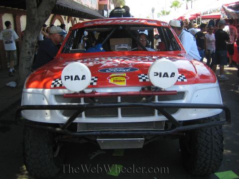 1990 Ford Bronco - Front view of a red-and-white Ford off-road race truck numbered 229 with twin PIAA lights, surrounded by spectators in the shade.