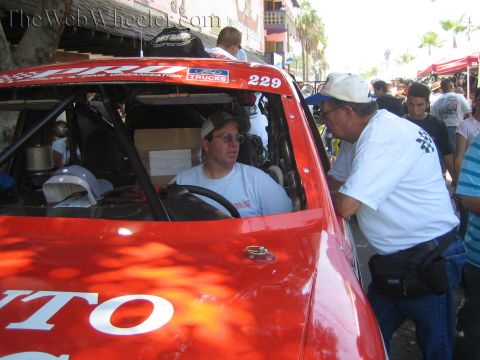 1990 Ford Bronco - driver in a white cap sits inside red off-road race truck 229 while another man in a cap leans toward the open window to talk.