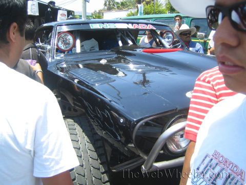 1990 Ford Bronco - black off-road race vehicle marked Ensenada with flame graphics and a roll cage is surrounded by onlookers in a sunny street.