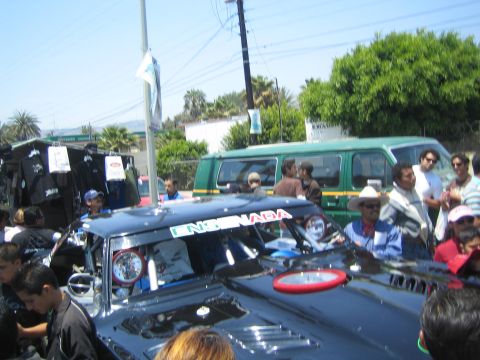 1990 Ford Bronco - dark Ensenada-marked off-road race car sits in a crowded street with a green van and palm trees behind it under a clear sky.