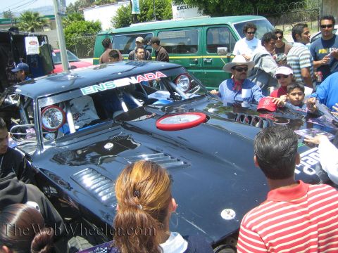 1990 Ford Bronco - Spectators crowd around a black off-road race car with an Ensenada windshield banner and a green van parked behind.