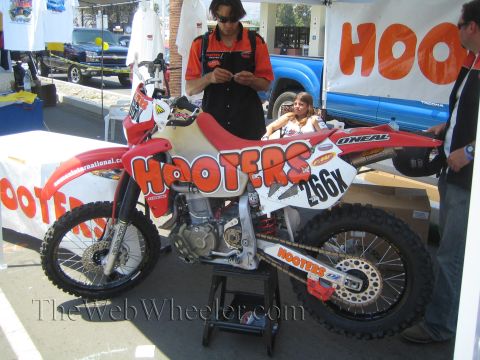 1990 Ford Bronco - red-and-white Hooters-sponsored dirt bike numbered 266X sits on a stand at a vendor booth as a person fills out paperwork.