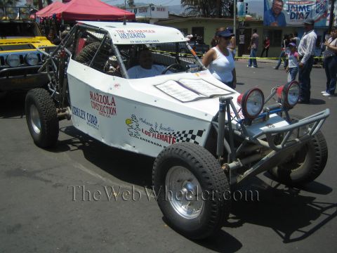 1990 Ford Bronco - white single-seat tube-frame off-road buggy lettered Marzola Construction and Kirkley Corp rolls along a sunny street.