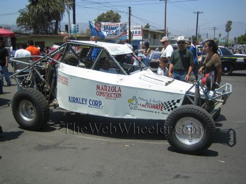 1990 Ford Bronco - Side view of a white tube-frame off-road race buggy marked Marzola Construction and Kirkley Corp on a street lined with spectators.