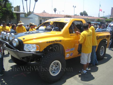 1990 Ford Bronco - yellow off-road race truck with Goodyear and Green branding is tended by a crew in matching yellow shirts on a street.