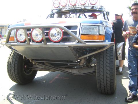 1990 Ford Bronco - Front low-angle view of a blue off-road race truck with a tube bumper, three round driving lights, and a roof light bar.