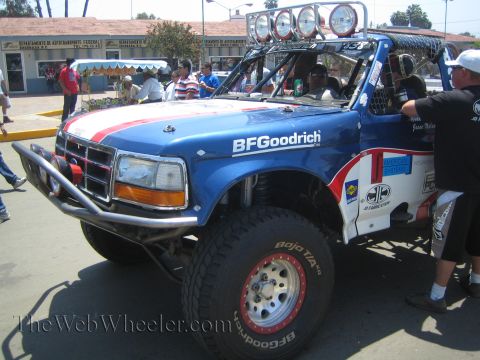 1990 Ford Bronco - blue Ford off-road race truck with a red-and-white stripe, BFGoodrich tires, and a tube bumper parked on a street.
