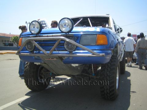 1990 Ford Bronco - Front view of a blue off-road race truck with a polished tube bumper, four round driving lights, and big off-road tires.