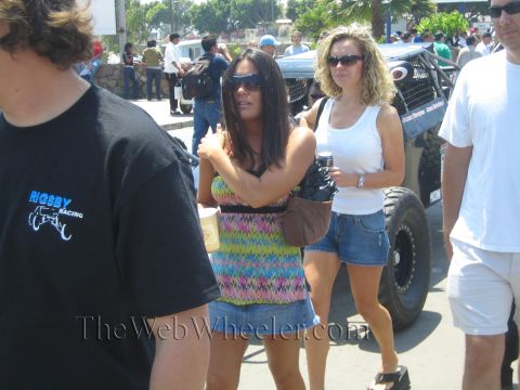 1990 Ford Bronco - Two women in sunglasses and casual summer clothes walk through a crowd past a parked off-road race car on a sunny day.