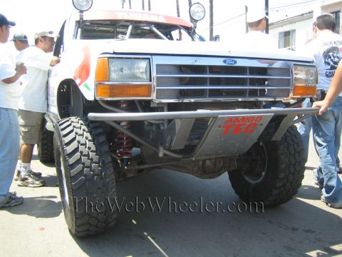 1990 Ford Bronco - Front close-up of a white Ford off-road truck showing its chrome grille, tube bumper, coilover suspension, and Amigo Tec decal.