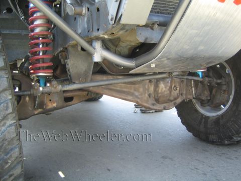 1990 Ford Bronco - Underside view of an off-road vehicle showing a solid front axle, red coilover shock, steering linkage, and tube frame.