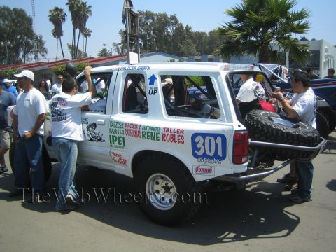 1990 Ford Bronco - white off-road SUV numbered 301 with a rear spare tire and Spanish sponsor lettering is surrounded by people on a street.
