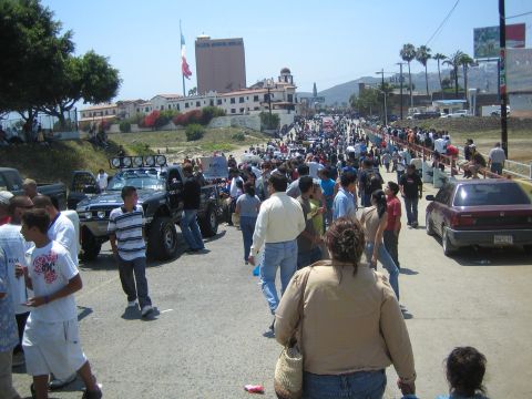 1990 Ford Bronco - wide street packed with pedestrians and off-road trucks, with a Mexican flag and hillside buildings in the background.
