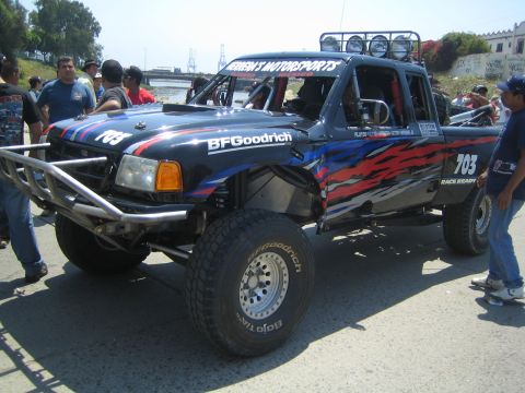 1990 Ford Bronco - black off-road prerunner race truck numbered 703 with BFGoodrich tires, tube bumpers and roof lights sits parked among a crowd.
