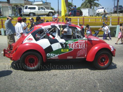 1990 Ford Bronco - red and white Volkswagen Beetle Baja race car numbered 1100 with Tecate sponsorship sits in a Mexican street amid spectators.