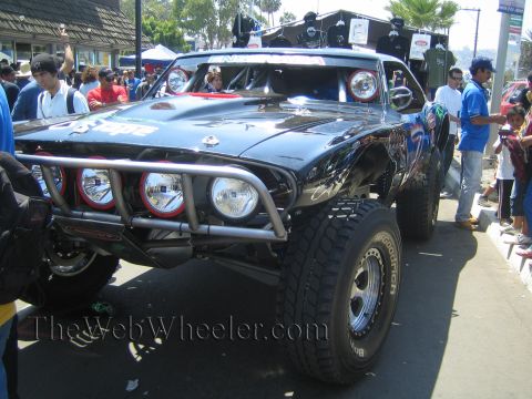 1990 Ford Bronco - glossy black off-road race vehicle with four round driving lights and a tube bumper sits surrounded by a street crowd.