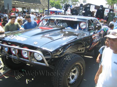 1990 Ford Bronco - glossy black off-road race car with roof-mounted lights and a tube bumper draws onlookers beside merchandise tents.