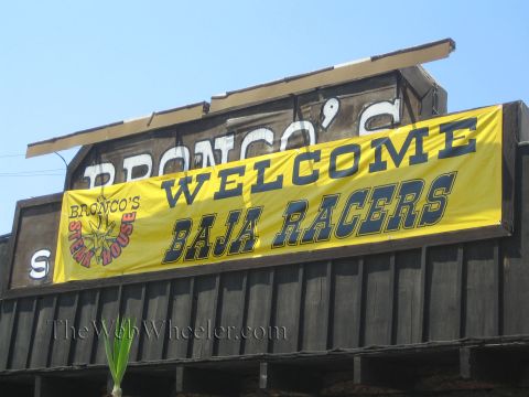 1990 Ford Bronco - yellow banner reading Welcome Baja Racers hangs below a painted Bronco's Steak House facade against a blue sky.
