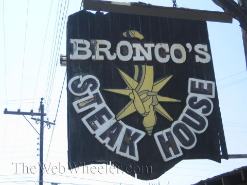 1990 Ford Bronco - weathered hanging sign reading Bronco's Steak House with a star-and-torch emblem is silhouetted against power lines and sky.