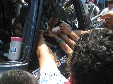 1990 Ford Bronco - Many hands reach into an open race truck door toward the driver as fans crowd around the vehicle on a sunny street.