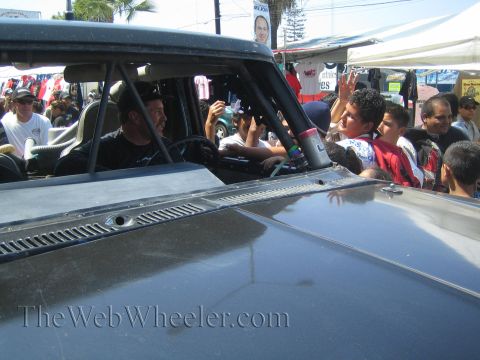 1990 Ford Bronco - Two men sit in a race truck cab waving to spectators lining a sunlit street full of vendor stalls and banners.