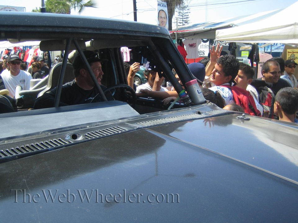 Two men seated in a race truck cab waving to a crowd along a street