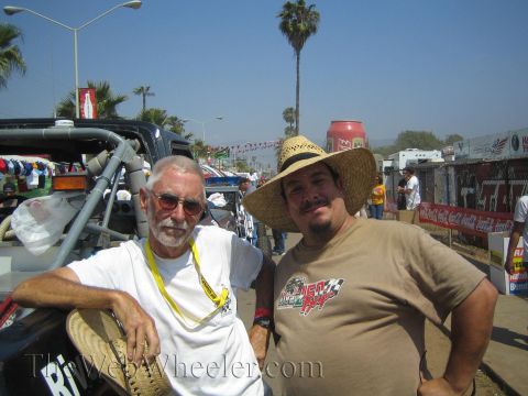 1990 Ford Bronco - older man with a white beard and a younger man in a straw hat pose together at an outdoor event with palm trees behind.