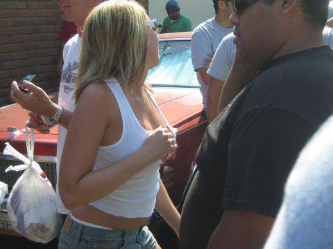 1990 Ford Bronco - blonde woman in a white tank top stands amid a street crowd beside a red sedan, holding a phone with onlookers around her.
