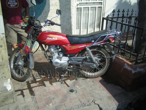 1990 Ford Bronco - red Italika TX150 motorcycle stands parked on a brick sidewalk beside a wrought-iron fence with a man standing behind it.