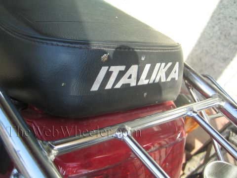 1990 Ford Bronco - close-up shows a black motorcycle seat printed ITALIKA in white above a chrome luggage rack and red bodywork.