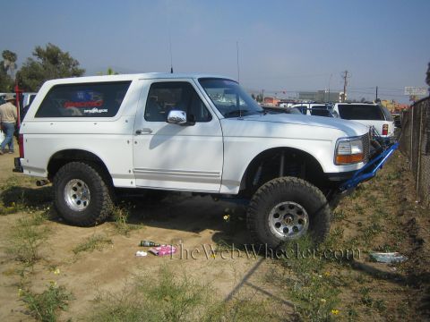 1990 Ford Bronco - white lifted Ford Bronco with a blue tube bumper and oversized tires sits parked in a dusty lot among other vehicles.